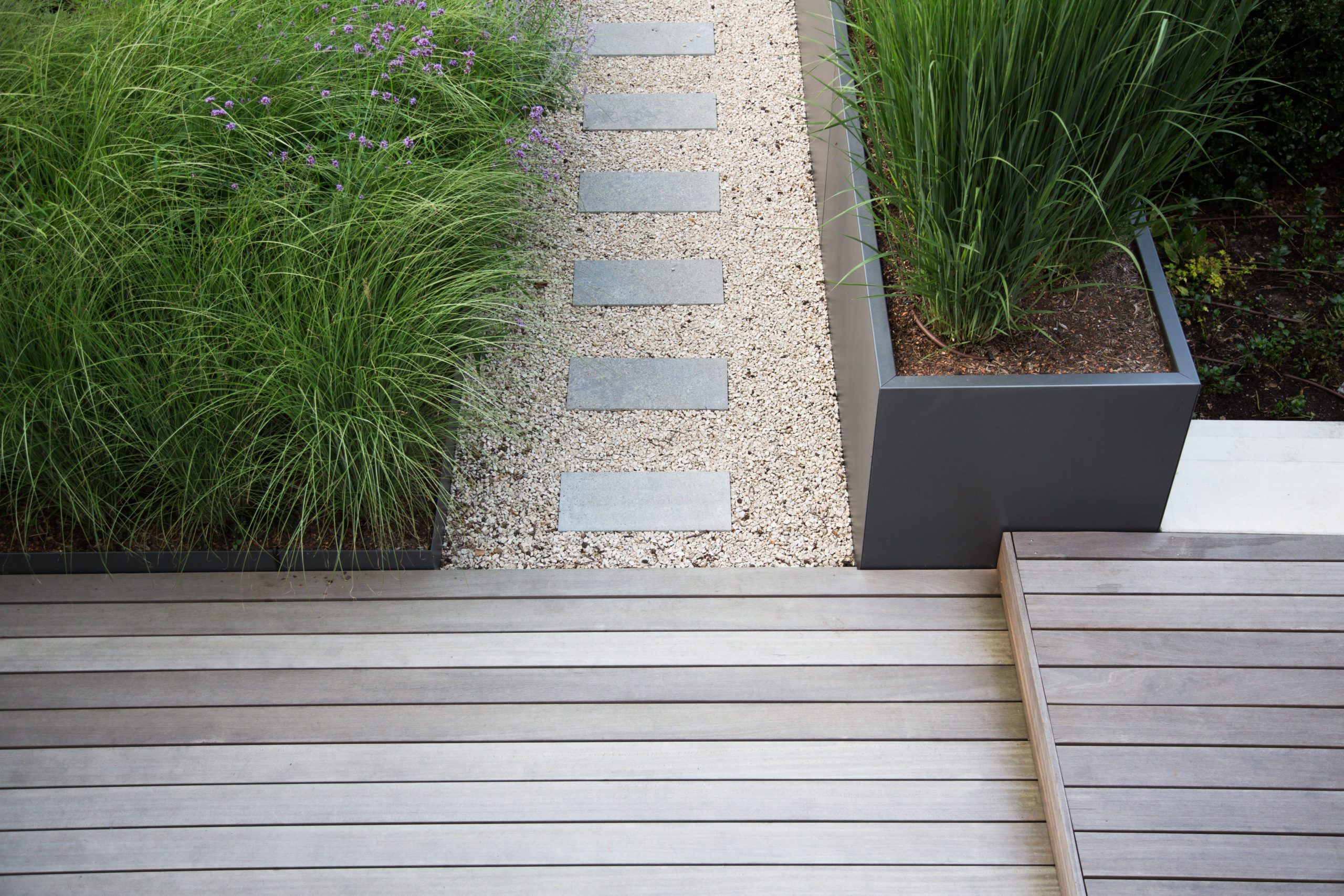 Moderne Garten- und Terrassengestaltung im Materialmix: Terrassen aus Holz und Gehweg aus Steinplatten umgeben von Schotter und Metall Pflanzgefäßen mit Grünpflanzen